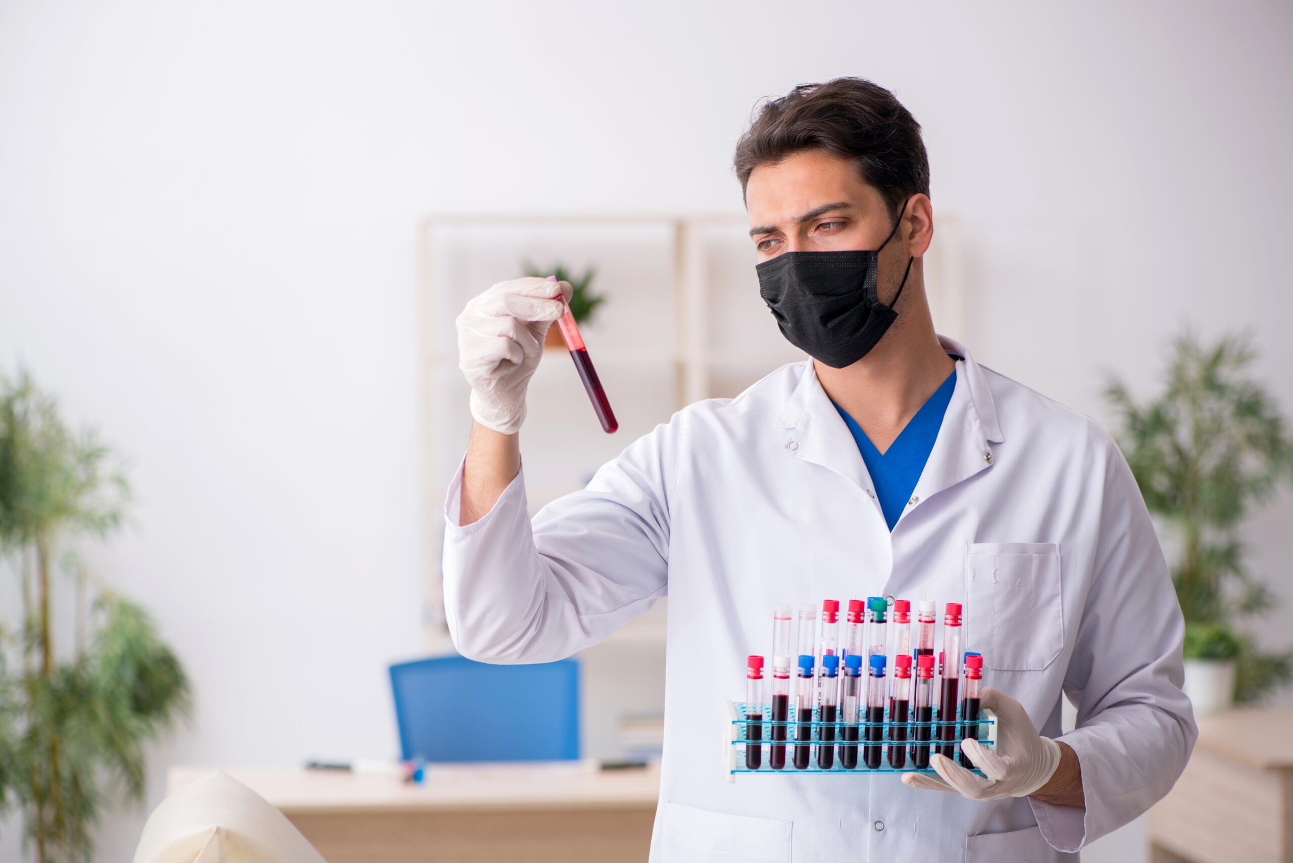 Um profissional de saúde, usando máscara preta e luvas, segura uma bandeja com tubos de ensaio cheios de sangue e observa atentamente um dos tubos. Um profissional de saúde, usando máscara preta e luvas, segura uma bandeja com tubos de ensaio cheios de sangue e observa atentamente um dos tubos.