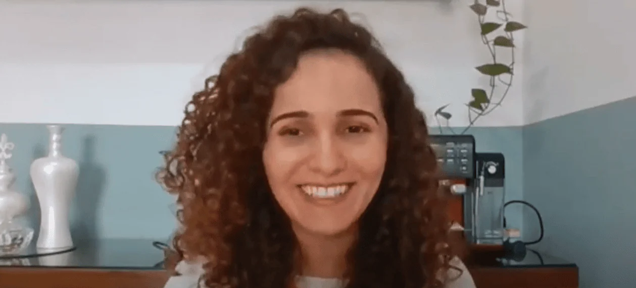 Suzana Suzana, palestrante, de cabelos cacheados, sorrindo, em fundo branco e cinza, com vasos de plantas e uma cafeteira