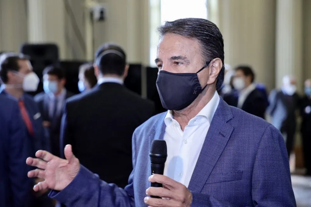 Jorge Moll de máscará preta, terno azul e blusa branca fala ao microfone durante evento. Atrás outras pessoas de máscara