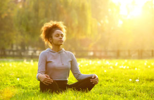 mulher negra meditando na grama com sol ameno no céu. Ela está de blusa cinza e calça preta. A meditação pode ser uma boa solução para se conhecer melhor e viver a sexualidade plenamente