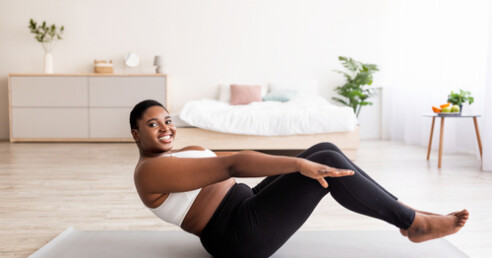 mulher negra acima do peso, de blusa branca e calça preta se exercita na sala de casa