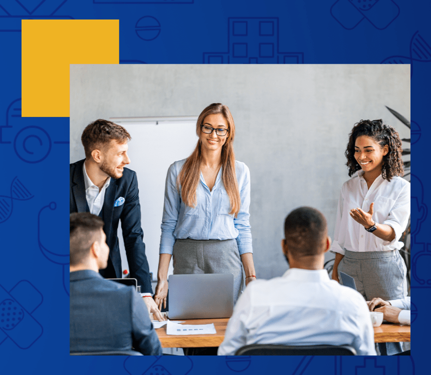 Cinco jovens profissionais ao redor de uma mesa de reunião, três estão de pé: um homem e duas mulheres e dois sentados: 2 homens. Dois dos homens usa terno e os demais blusas sociais. Parecem conversar e estão sorrindo