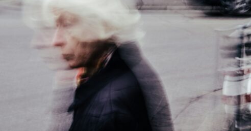 Imagem mostra mulher idosa de perfil, atravessando a rua, com casaco de frio preto, e tem um padrão estético em movimento, mexido, borrado, como se o fotógrafo tivesse mexido a câmera no momento da foto.