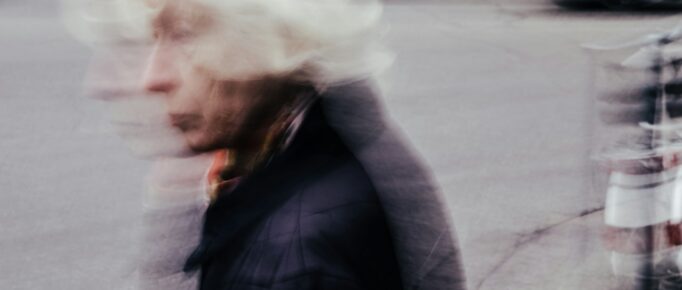 Imagem mostra mulher idosa de perfil, atravessando a rua, com casaco de frio preto, e tem um padrão estético em movimento, mexido, borrado, como se o fotógrafo tivesse mexido a câmera no momento da foto.