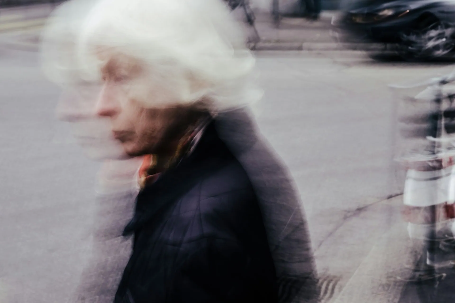 Imagem mostra mulher idosa de perfil, atravessando a rua, com casaco de frio preto, e tem um padrão estético em movimento, mexido, borrado, como se o fotógrafo tivesse mexido a câmera no momento da foto.