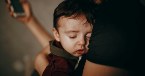 Mãe de blusa preta segurando um bebê num braço e no outro segurando um celular e fazendo uma ligação.