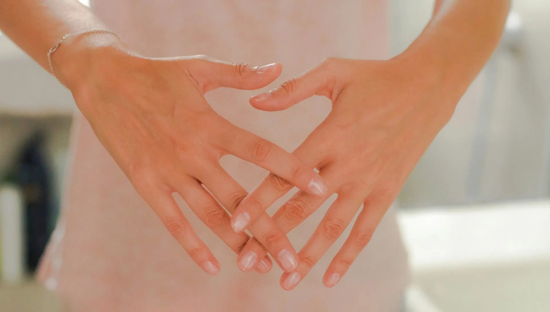 ALT: imagem em tom pastel mostra uma mulher em pé, com seu corpo em segundo plano, e em primeiro plano suas duas mãos com os dedos entrelaçados, à frente do corpo, na altura da região pélvica.