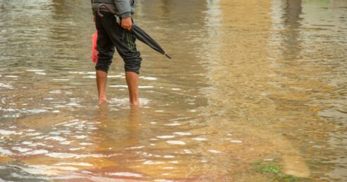 ALT: imagem mostra um homem segurando um guarda-chuva fechado, atravessando uma rua alagada, descalço, com a calça puxada até o joelho.