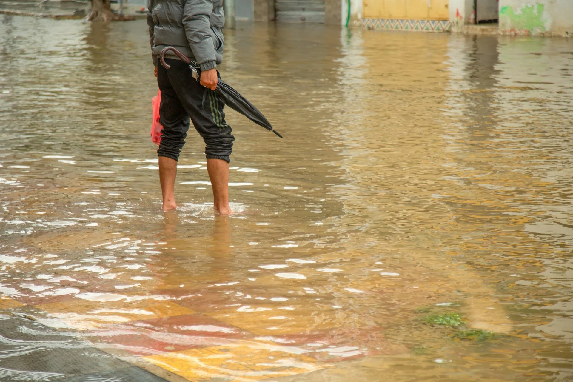 ALT: imagem mostra um homem segurando um guarda-chuva fechado, atravessando uma rua alagada, descalço, com a calça puxada até o joelho.