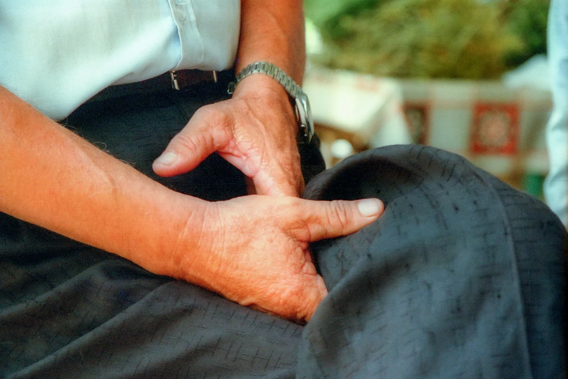 ALT: imagem mostra um homem de calça cinza, camisa branca e cinto preto, sentado de penas cruzadas, com as mãos na altura da virilha.