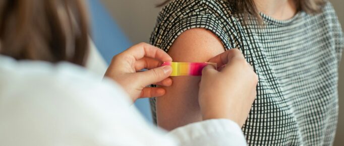 ALT: imagem mostra em primeiro plano uma profissional da saúde colocando um bandaid no braço da paciente, em segundo plano, que acabou de tomar uma vacina.