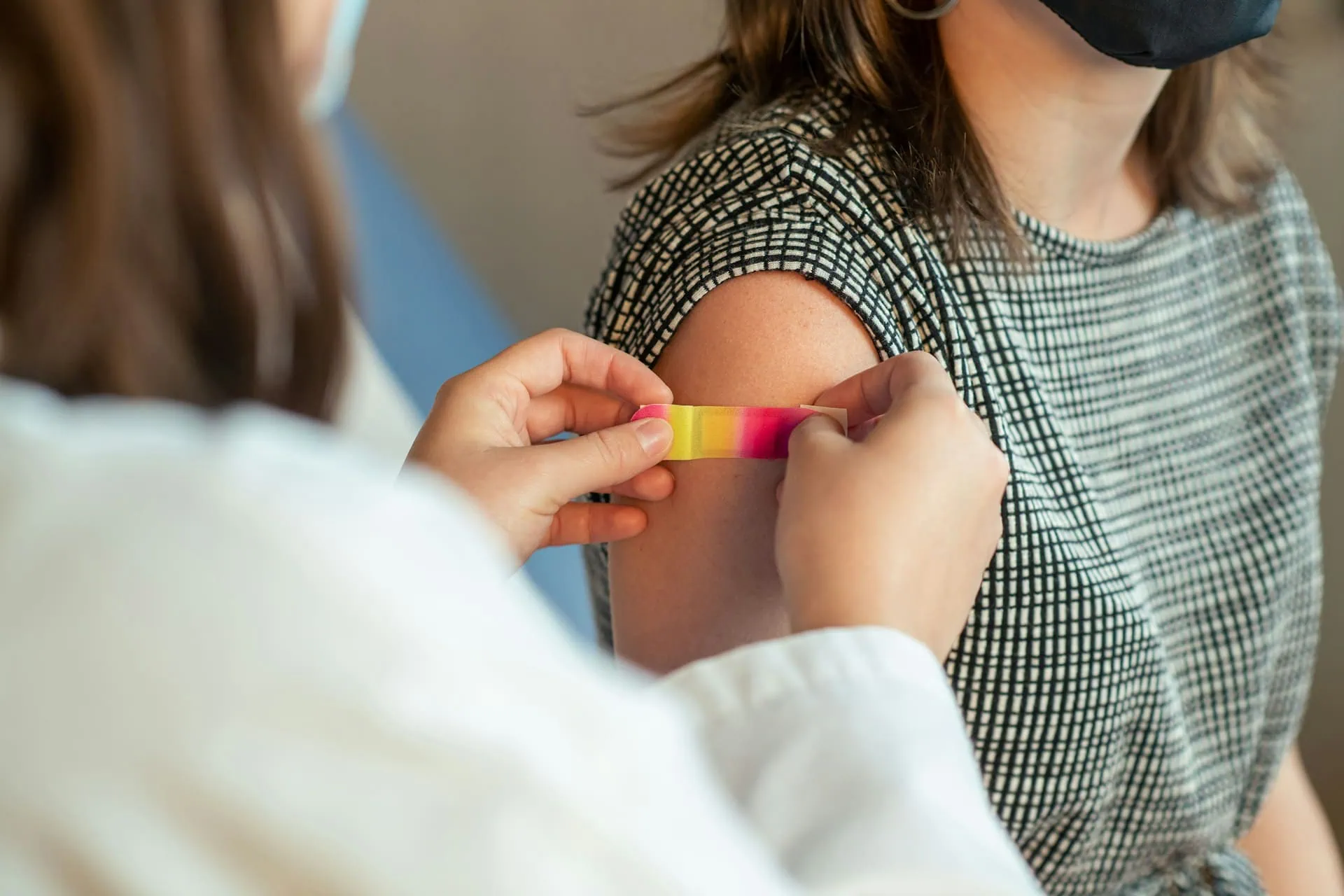 ALT: imagem mostra em primeiro plano uma profissional da saúde colocando um bandaid no braço da paciente, em segundo plano, que acabou de tomar uma vacina.