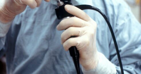 ALT: imagem mostra um médico, usando óculos de grau de armação preta e vestimentas azuis utilizadas em centros cirúrgicos, olhando através de um colonoscópio (parte do instrumento usado na colonoscopia).