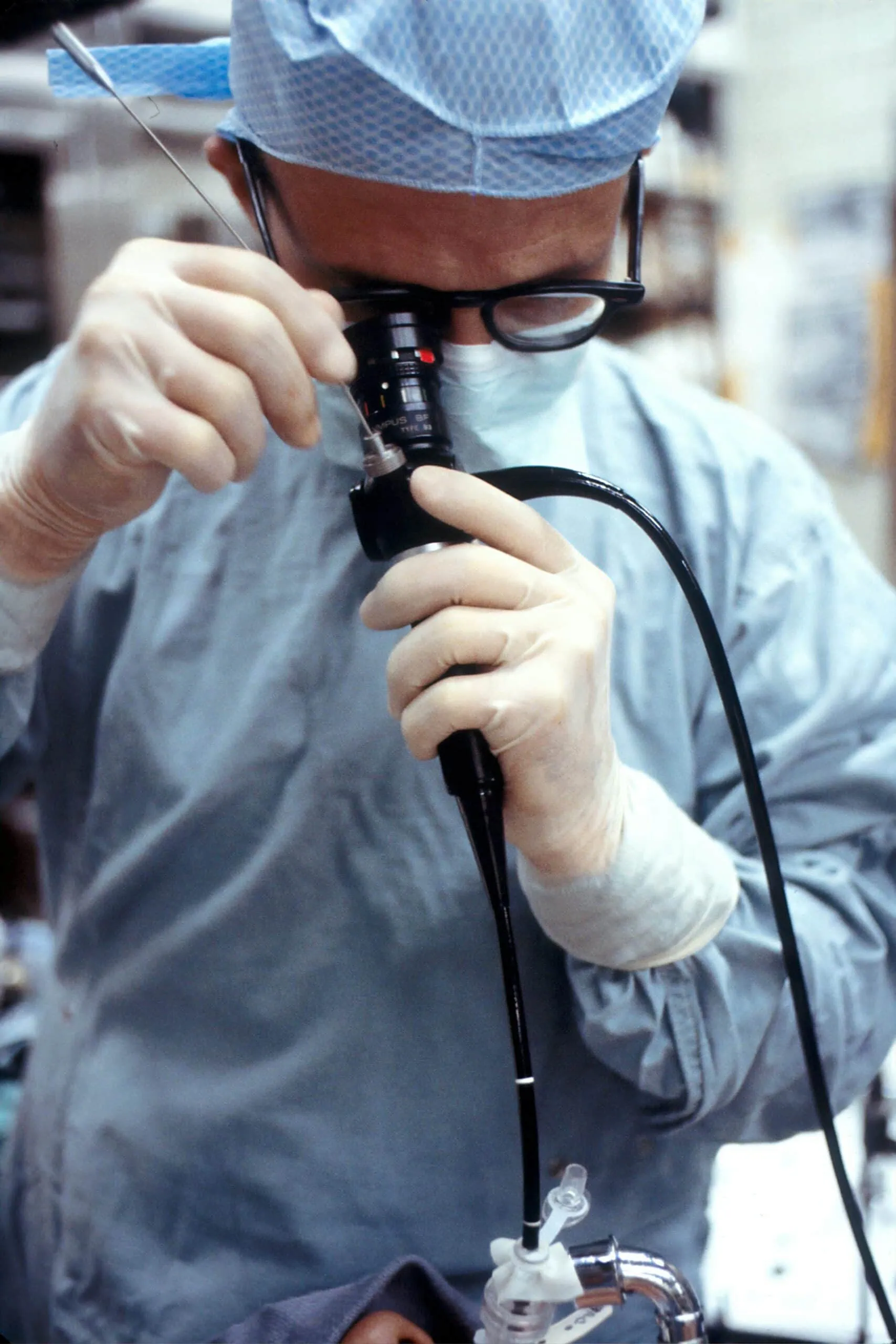 ALT: imagem mostra um médico, usando óculos de grau de armação preta e vestimentas azuis utilizadas em centros cirúrgicos, olhando através de um colonoscópio (parte do instrumento usado na colonoscopia).