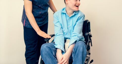 ALT: criança sorridente, de pele branca e cabelo castanho curto, de blusa azul e calça jeans, sentada em cadeira de rodas, e acompanhada por enfermeira de blusa e calça azul escura.
