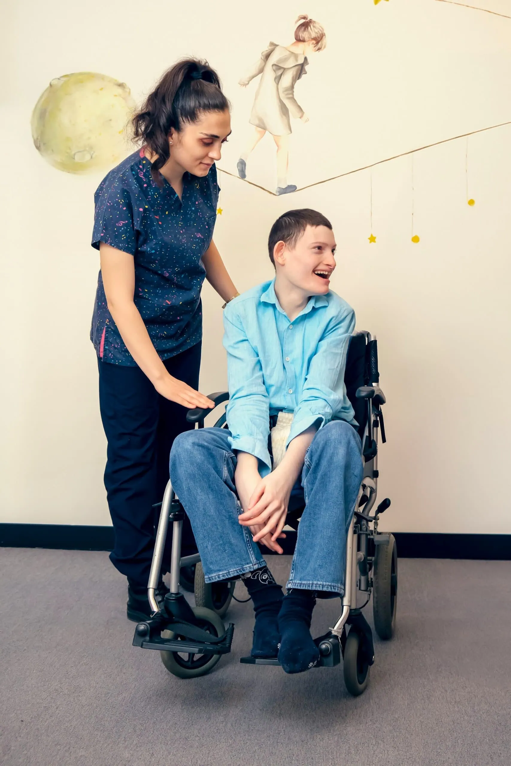 ortopediatri-cocuk-ortopedi-akademisi-alCU30xHRnA-unsplash ALT: criança sorridente, de pele branca e cabelo castanho curto, de blusa azul e calça jeans, sentada em cadeira de rodas, e acompanhada por enfermeira de blusa e calça azul escura.