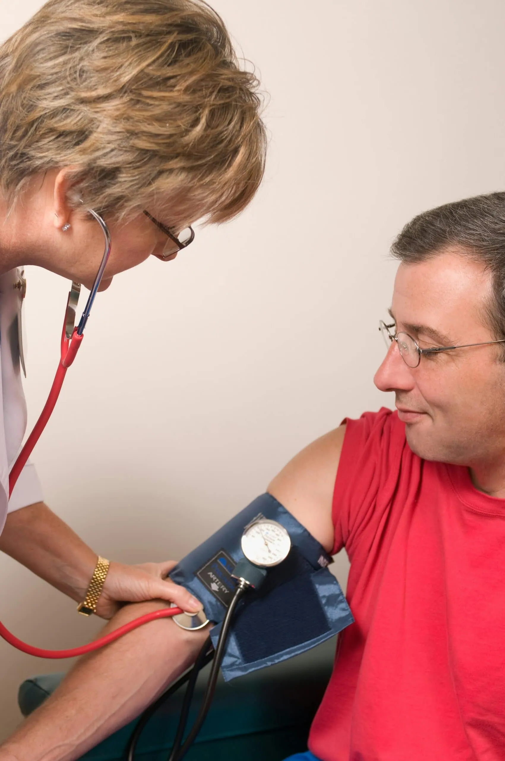saúde-do-homem-unsplash ALT: imagem mostra médica de jaleco branco, segurando estetoscopio e medindo a pressão de um homem de meia-idade de óculos e camiseta vermelha.
