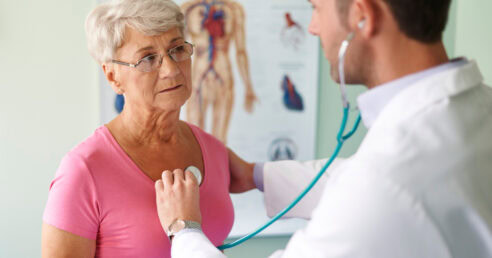 ALT: mulher numa consulta médica, sendo examinada por um médico que está auscultando e checando os batimentos cardíacos da paciente.