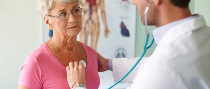 ALT: mulher numa consulta médica, sendo examinada por um médico que está auscultando e checando os batimentos cardíacos da paciente.