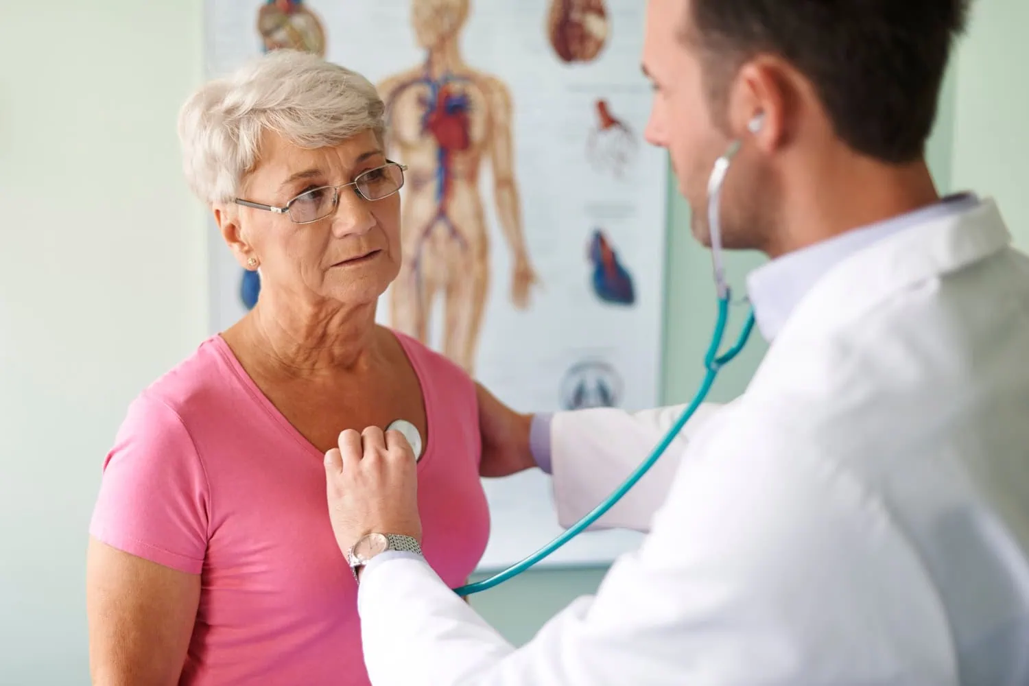 ALT: mulher numa consulta médica, sendo examinada por um médico que está auscultando e checando os batimentos cardíacos da paciente.