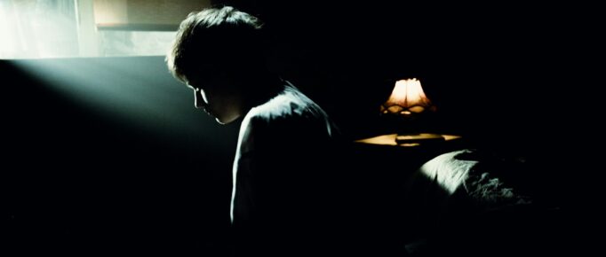 ALT: homem sentado numa cama, de cabeça baixa, visto de lado, em ambiente com pouca luz, que sugere ser o quarto de dormir.
