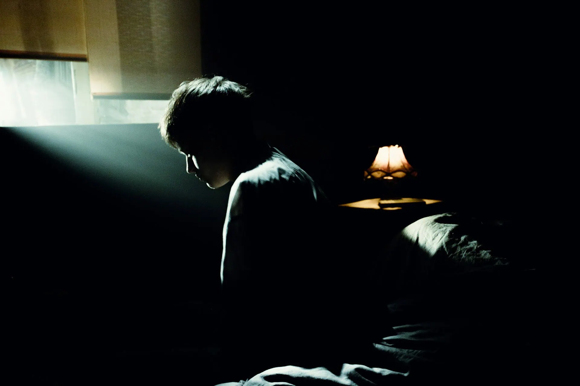 ALT: homem sentado numa cama, de cabeça baixa, visto de lado, em ambiente com pouca luz, que sugere ser o quarto de dormir.