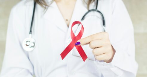 ALT: médica vestindo jaleco branco, com estetoscópio no pescoço, segura fita vermelho símbolo da conscientização e luta contra a aids e hiv.
