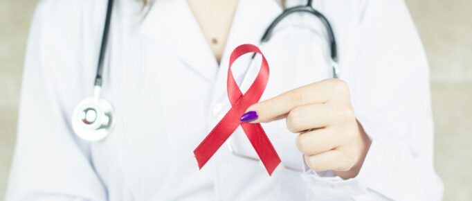 ALT: médica vestindo jaleco branco, com estetoscópio no pescoço, segura fita vermelho símbolo da conscientização e luta contra a aids e hiv.