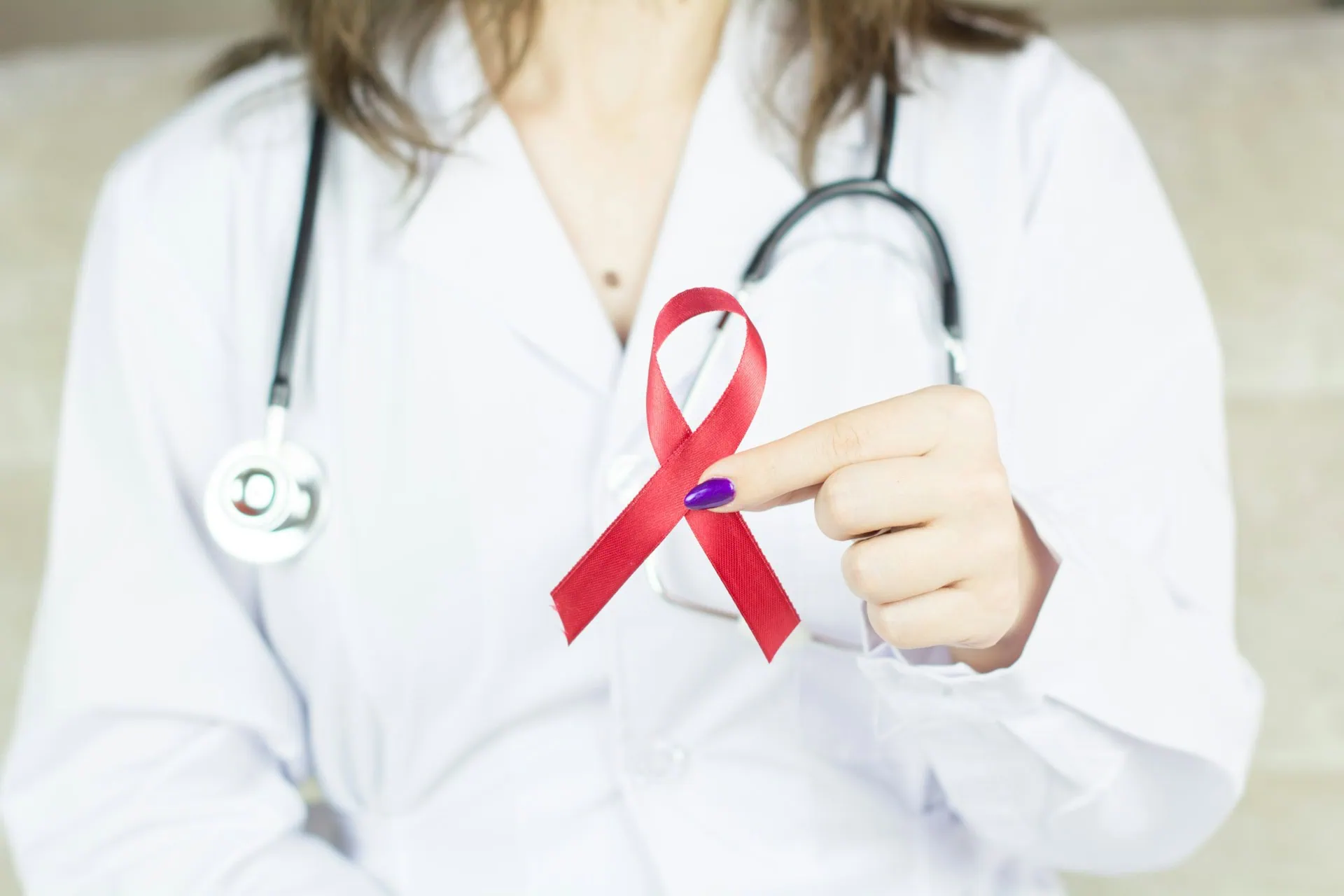 bermix-studio-Gg6OrxUotLQ-unsplash ALT: médica vestindo jaleco branco, com estetoscópio no pescoço, segura fita vermelho símbolo da conscientização e luta contra a aids e hiv.