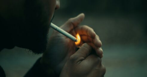 ALT: close do rosto de um homem, de lado, com pele branca e barba curta, acendendo um cigarro.