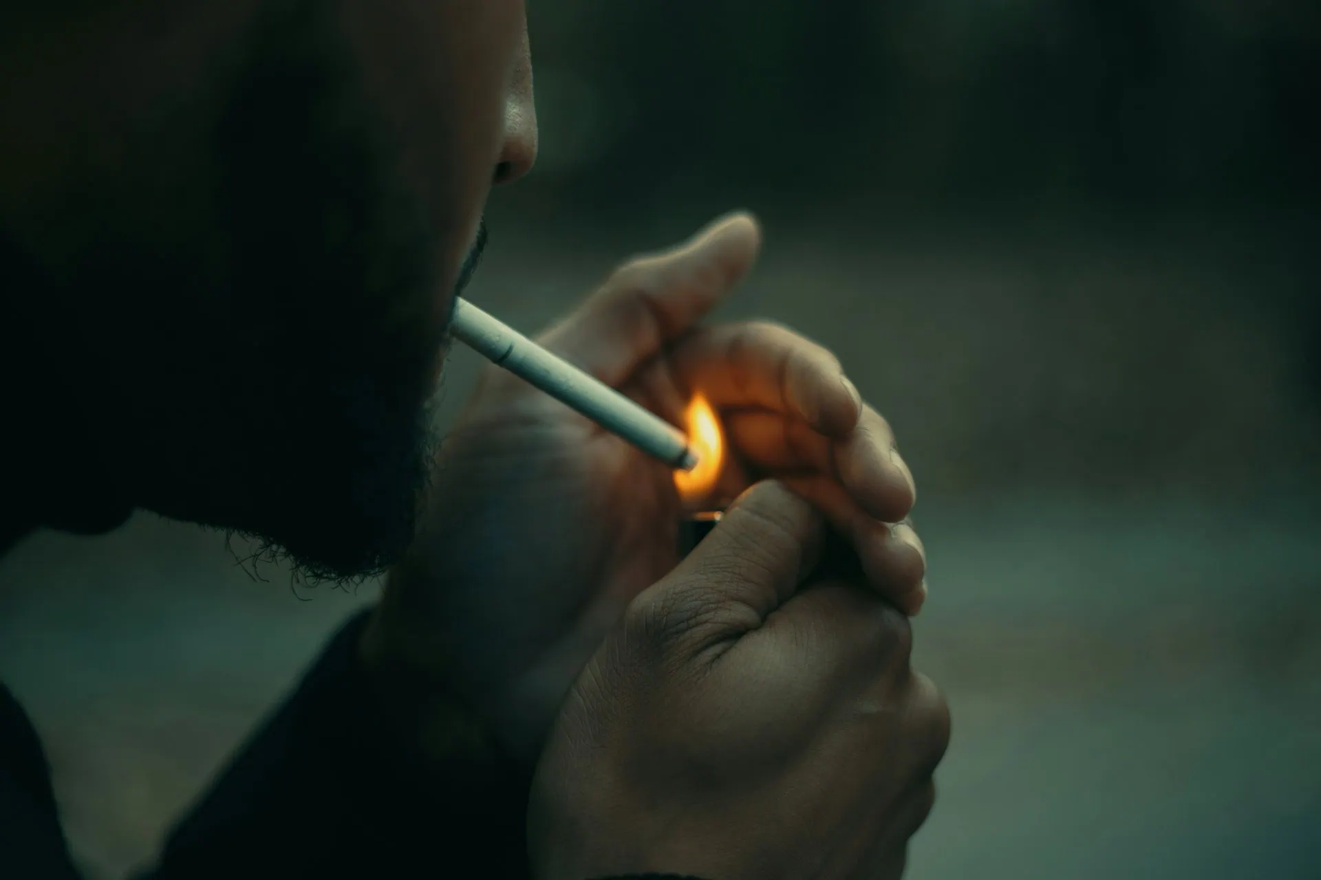ALT: close do rosto de um homem, de lado, com pele branca e barba curta, acendendo um cigarro.