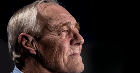 ALT: sob fundo preta, aparece o perfil de homem de idade avançada, cabelo grisalho, pele branca e olhos claro, com olhar longe e perdido.
