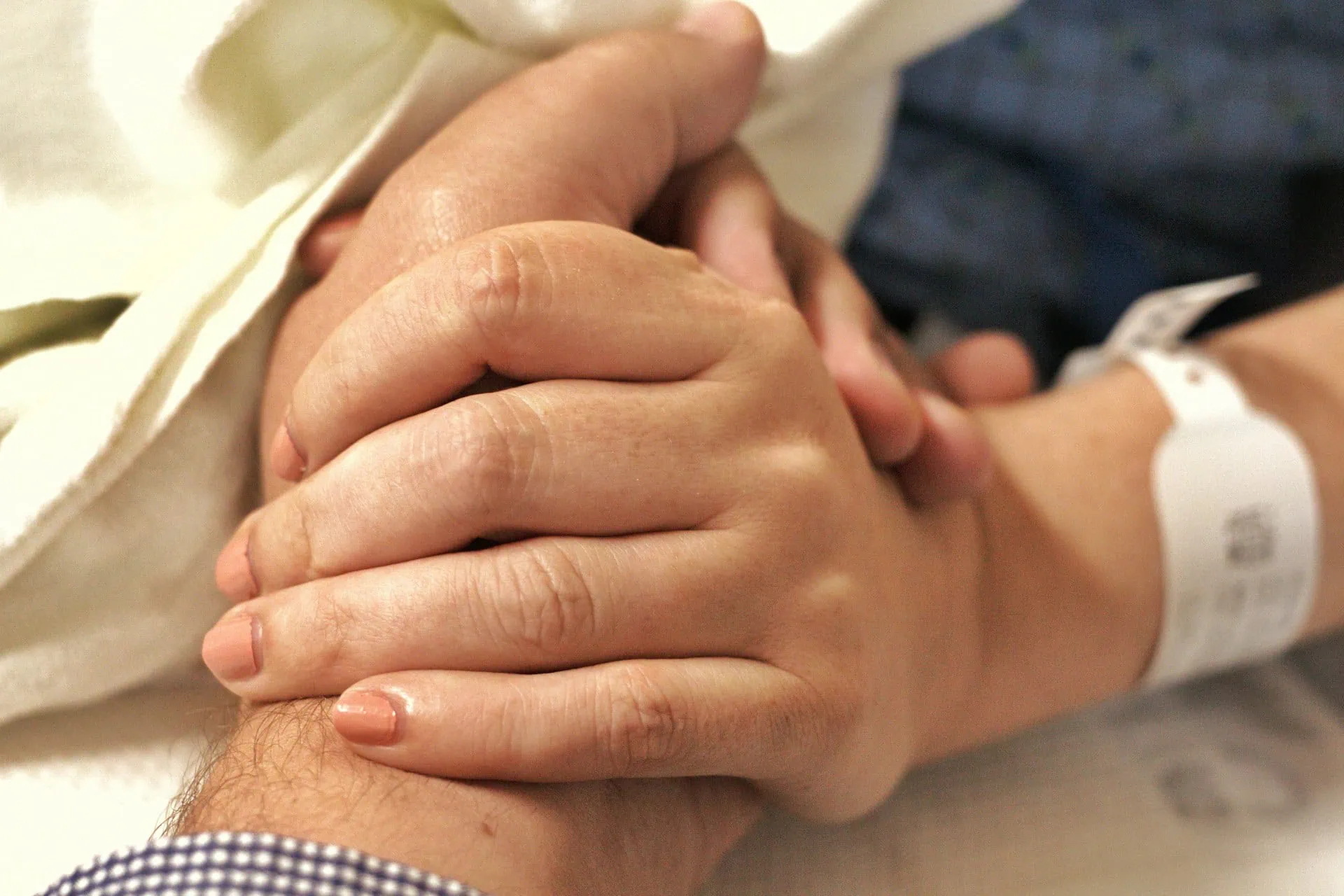 ALT: imagem em primeiro plano de duas mãos dadas, ambientado em cama de hospital, onde um dos braços mostram pulseira de internação hospitalar.