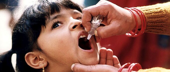 ALT: Uma menina pequena, de cabelos e olhos escuros, recebe dose oral da vacina contra a poliomielite em formato de gotinhas, ministrada por profissional da saúde.