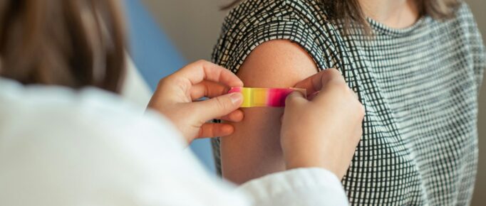 ALT: imagem recortada, foco na altura do ombro de uma mulher, pele branca, que acabou de ser vacinada e recebe de&nbsp;uma enfermeira&nbsp;um curativo do tipo band-aid no braço.