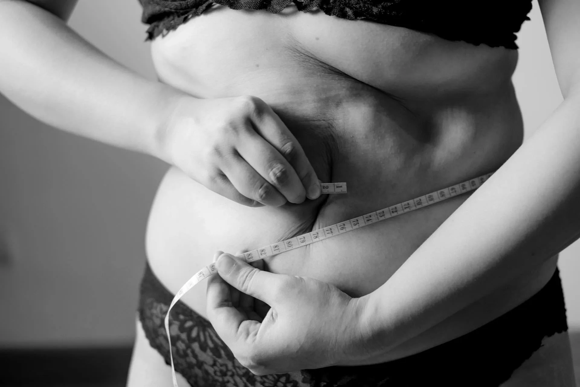 ALT: mulher vestida apenas com o sutiã, cujo sobrepeso pode ser visualizado pela gordura na barriga, tenta medir a cintura com fita métrica.