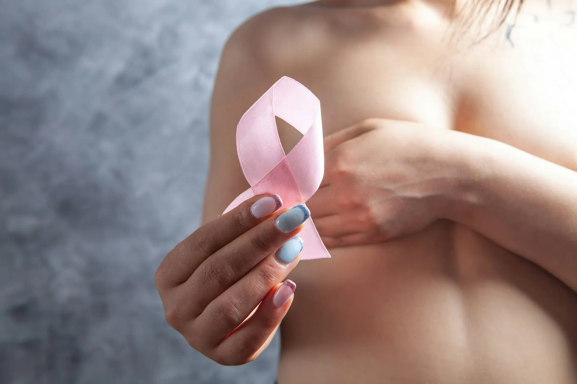 ALT: Foto recortada na altura dos seios de mulher branca, com o colo desnudo, que esconde os seios com uma mão, e na outra a fita rosa símbolo da conscientização sobre o câncer de mama.
